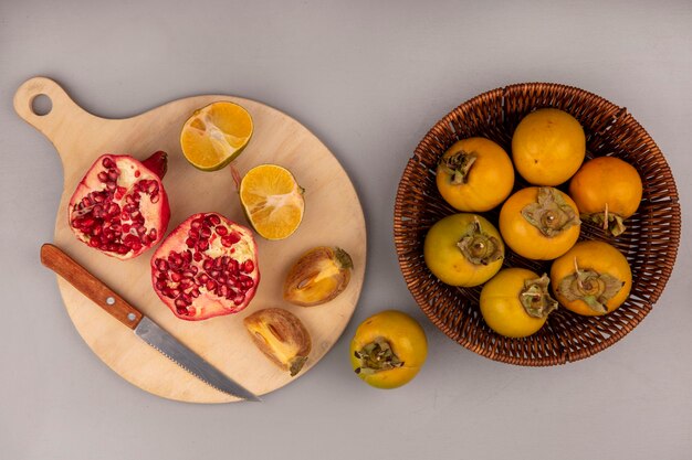 Vue de dessus de la grenade fraîche coupée en deux sur une planche de cuisine en bois avec un couteau avec des fruits kaki coupés en deux et des mandarines isolées