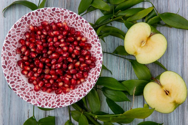 Vue de dessus de la grenade pelée sur une assiette avec les moitiés d'une pomme verte et des branches de feuilles sur une surface grise