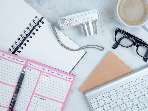 Vue de dessus en gros plan du cahier, du planificateur, des lunettes, du clavier, du café et de l'appareil photo sur le lit