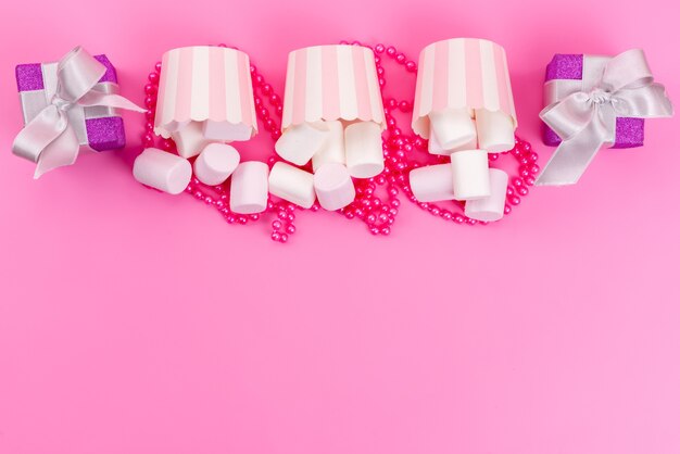 Une vue de dessus des guimauves blanches à l'intérieur des emballages en papier avec de petites boîtes-cadeaux violettes sur bureau rose, gâteau meringue sucré