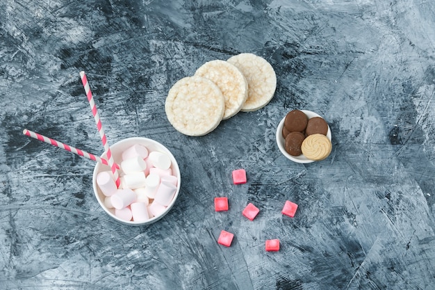 Vue de dessus des guimauves et des cannes à sucre dans une tasse avec des gaufrettes de riz, des biscuits et des bonbons sur une surface en marbre bleu foncé. horizontal