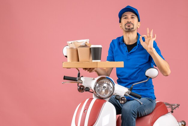 Vue de dessus de l'homme confiant de messagerie portant un chapeau assis sur un scooter faisant un geste parfait sur fond de pêche pastel