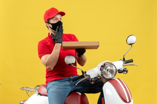 Vue de dessus de l'homme de messagerie choqué portant chemisier rouge et gants de chapeau en masque médical assis sur ordre de maintien de scooter