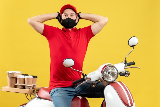 Vue de dessus de l'homme de messagerie confus portant un chemisier rouge et des gants de chapeau dans un masque médical délivrant l'ordre assis sur un scooter