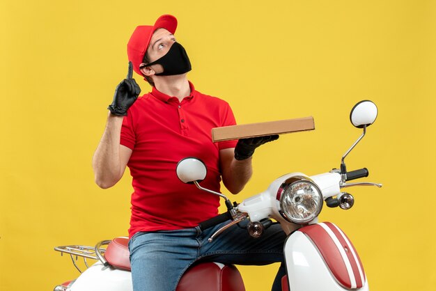 Vue de dessus de l'homme de messagerie confus portant chemisier rouge et gants chapeau en masque médical assis sur un scooter montrant l'ordre pointant vers le haut