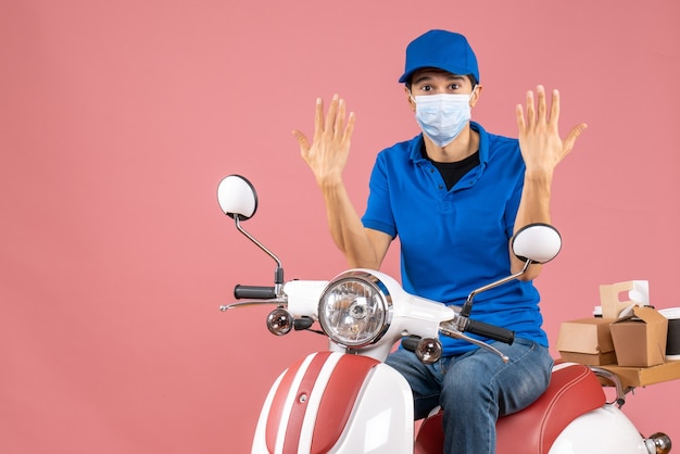 Vue de dessus de l'homme de messagerie dans un masque médical portant un chapeau assis sur un scooter se sentant curieux sur fond de pêche pastel