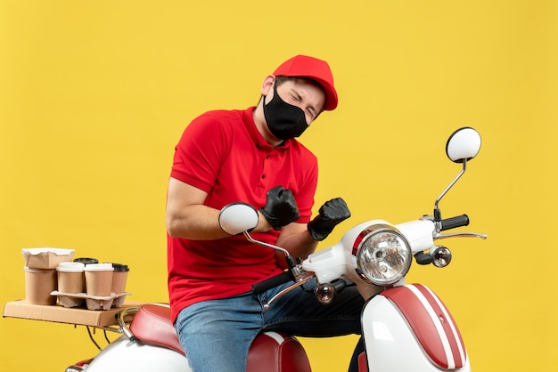 Vue de dessus de l'homme de messagerie heureux portant chemisier rouge et gants de chapeau en masque médical délivrant l'ordre assis sur scooter