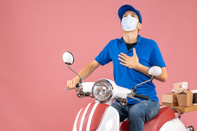 Vue de dessus de l'homme de messagerie en masque médical portant un chapeau assis sur un scooter se sentant fier sur fond de pêche pastel