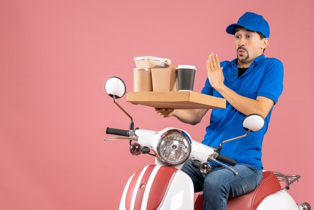 Vue de dessus d'un homme de messagerie paniqué portant un chapeau assis sur un scooter sur fond de pêche pastel