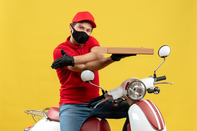 Vue de dessus de l'homme de messagerie souriant portant chemisier rouge et gants de chapeau en masque médical assis sur un scooter montrant l'ordre d'accueillir quelqu'un