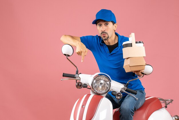 Vue de dessus de l'homme se demandant courrier portant un chapeau assis sur un scooter montrant des commandes pointant vers le bas sur une pêche pastel