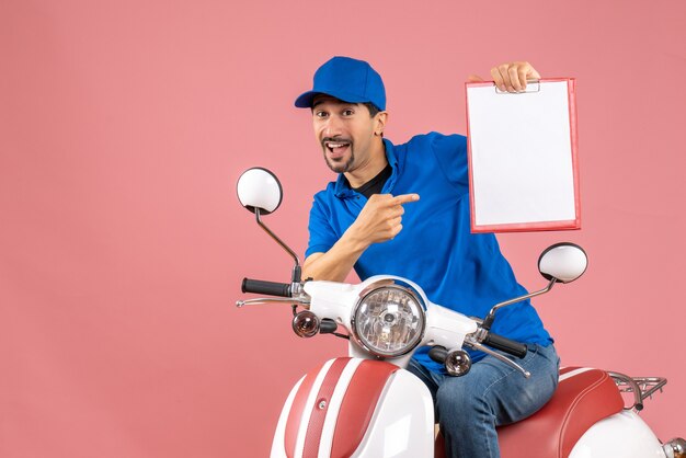 Vue de dessus d'un homme souriant portant un chapeau assis sur un scooter montrant un document sur une pêche pastel