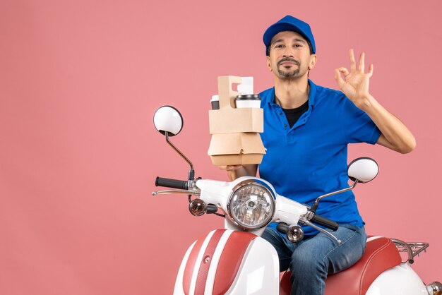 Vue de dessus d'un homme souriant portant un chapeau assis sur un scooter tenant des commandes faisant un geste parfait sur une pêche pastel