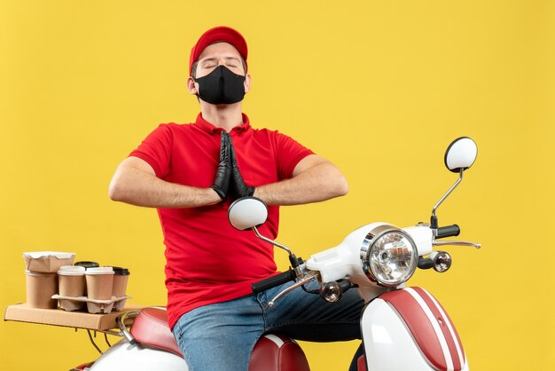 Vue de dessus d'un jeune adulte plein d'espoir portant un chemisier rouge et des gants de chapeau dans un masque médical délivrant l'ordre assis sur un scooter priant