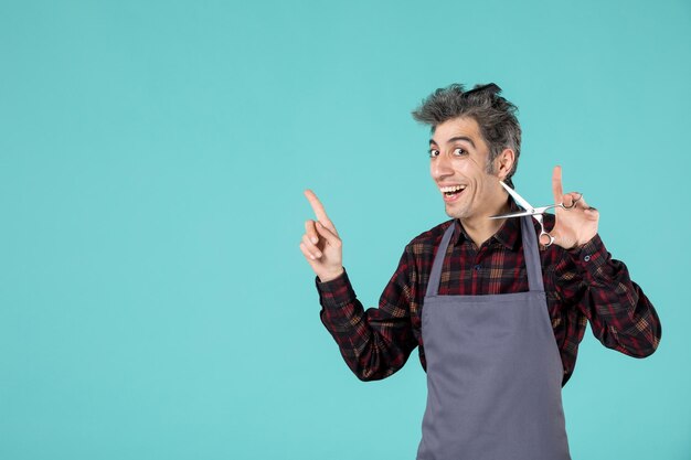 Vue de dessus d'un jeune coiffeur souriant portant un tablier gris et tenant un peigne sur la tête et montrant des ciseaux pointant quelque chose sur le côté droit sur fond bleu