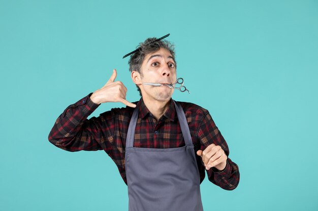 Vue de dessus d'un jeune coiffeur surpris portant un tablier gris et tenant un peigne sur la tête et gardant des ciseaux entre les lèvres en faisant un geste d'appel sur une surface de couleur bleu pastel