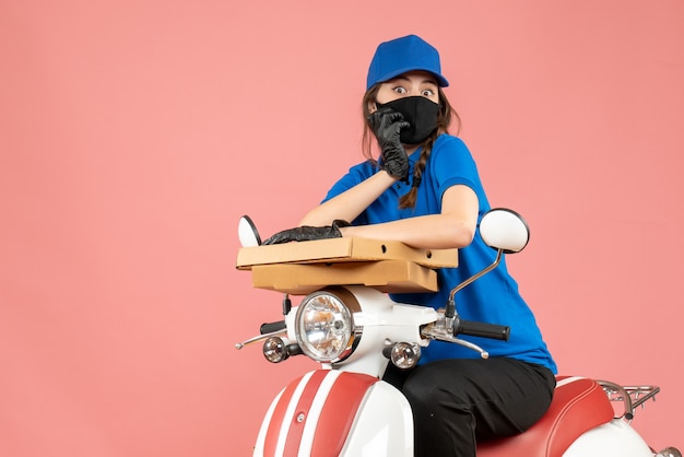 Vue de dessus d'une jeune coursière concernée portant un masque médical et des gants assis sur un scooter livrant des commandes sur une pêche pastel