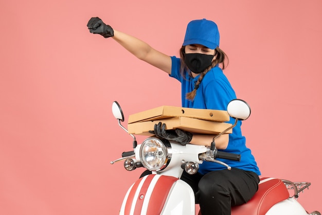 Vue de dessus d'une jeune coursière émotive et fière portant un masque médical et des gants assis sur un scooter livrant des commandes sur une pêche pastel