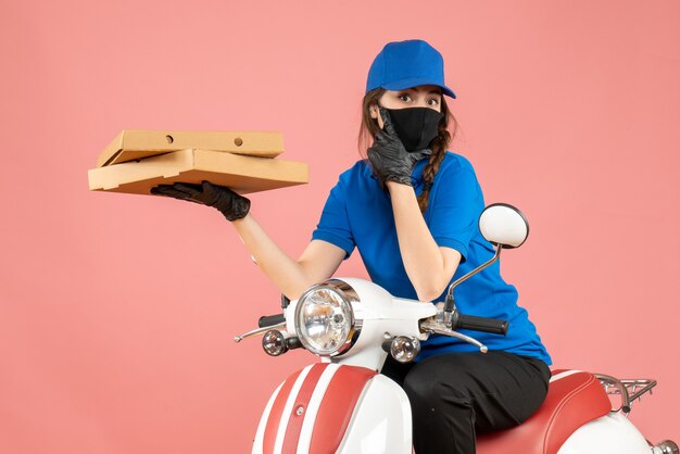 Vue de dessus d'une jeune coursière focalisée portant un masque médical et des gants assis sur un scooter livrant des commandes sur une pêche pastel