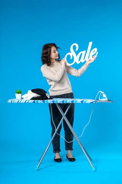 Vue de dessus d'une jeune femme confiante debout derrière la planche à repasser et montrant l'icône de vente sur fond bleu