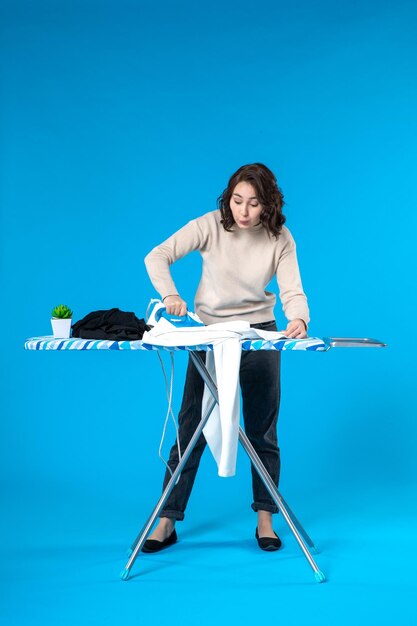 Vue de dessus d'une jeune femme curieuse debout derrière la planche et repassant les vêtements sur une surface bleue