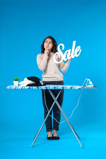 Vue de dessus d'une jeune femme réfléchie debout derrière la planche à repasser et montrant l'icône de vente sur fond bleu