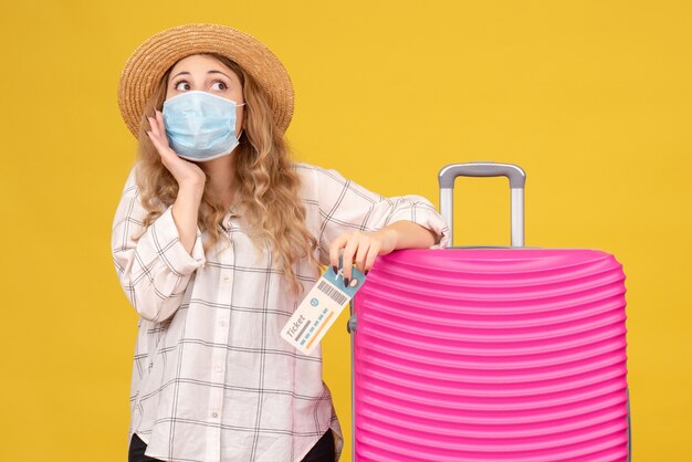 Vue de dessus d'une jeune femme réfléchie portant un masque montrant un billet et debout près de son sac rose