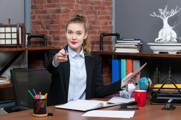 Vue de dessus d'une jeune femme souriante assise à une table et tenant un document pointant un stylo de couleur bleue au bureau