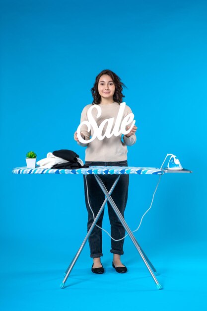 Vue de dessus d'une jeune femme souriante debout derrière la planche à repasser et montrant l'icône de vente sur fond bleu