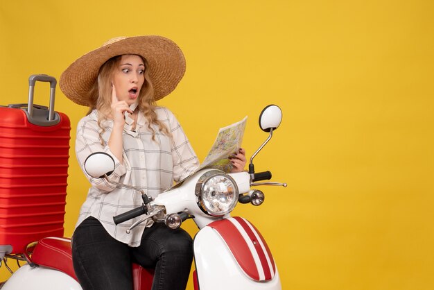 Vue de dessus de la jeune femme surprise portant un chapeau et assis sur la moto et regardant la carte pointant vers le haut sur jaune