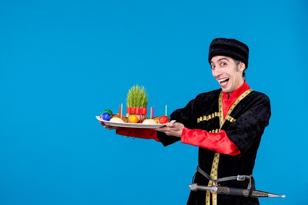 Vue de dessus d'un jeune homme souriant en costume traditionnel montrant un plateau rempli de confiseries nationales sur fond bleu