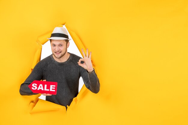 Vue de dessus d'un jeune homme souriant tenant une pancarte de vente et faisant un geste de lunettes dans un mur jaune déchiré