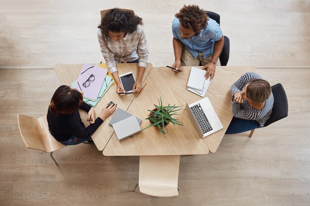 Vue de dessus de jeunes entrepreneurs professionnels du groupe assis à table dans l'espace de coworking, discutant des bénéfices du dernier projet d'équipe, à l'aide d'un ordinateur portable, d'une tablette numérique et d'un smartphone.