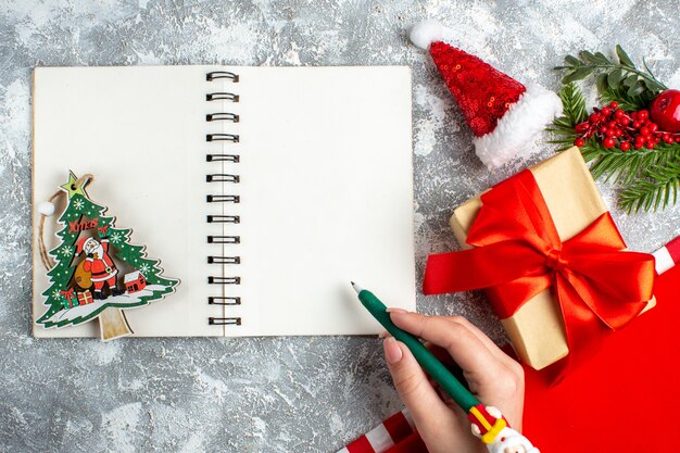 Vue de dessus jouet de Noël sur ordinateur portable petit bonnet de noel présent stylo dans une main féminine sur un tableau blanc gris