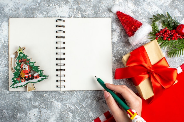 Vue de dessus jouet de Noël sur ordinateur portable petit bonnet de noel présent stylo dans une main féminine sur un tableau blanc gris