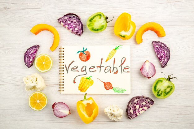 Vue de dessus légume écrit sur le bloc-notes légumes coupés chou rouge tomate verte citrouille oignon rouge poivron jaune chou-fleur citron sur tableau blanc