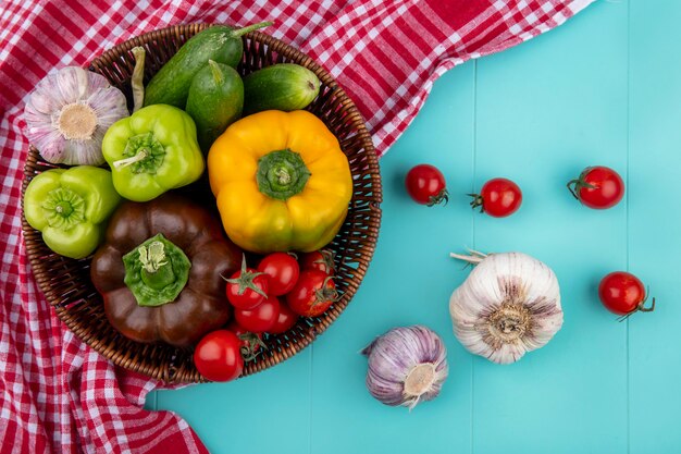 Vue de dessus des légumes comme poivron concombre tomate ail dans le panier sur tissu à carreaux et sur la surface bleue