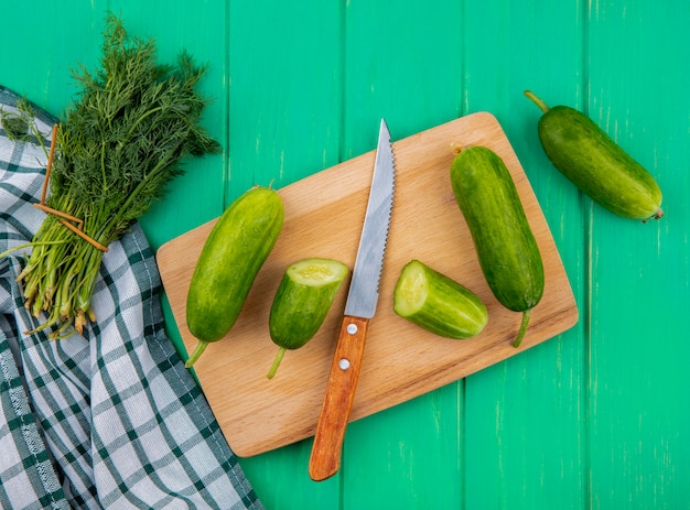 Vue de dessus des légumes coupés et concombre entier avec un couteau sur une planche à découper et un bouquet d'aneth sur un tissu à carreaux et une surface verte