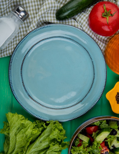 Vue de dessus des légumes coupés et entiers comme la laitue concombre tomate basilic avec sel poivre noir et assiette vide sur vert