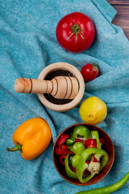 Vue de dessus des légumes entiers et des poivrons et des tomates en tranches avec du citron et du poivre noir dans un broyeur d'ail sur un tissu bleu