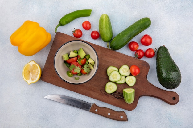 Vue de dessus de légumes frais comme concombre haché tomates cerises poivron jaune et citron sur planche de cuisine avec couteau sur blanc