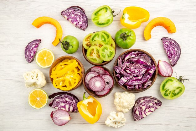 Vue de dessus légumes frais coupés chou rouge tomate verte citrouille oignon rouge poivron jaune chou-fleur citron dans de petits bols sur une surface en bois blanc