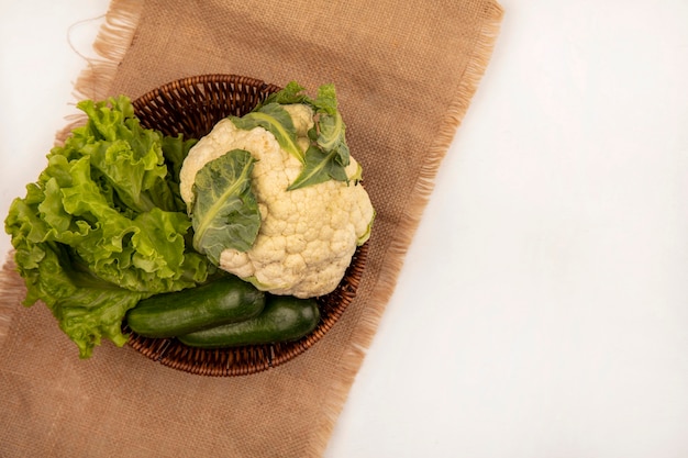 Vue de dessus de légumes frais tels que le chou-fleur de laitue et les concombres sur un seau sur un sac en tissu sur un fond blanc avec copie espace