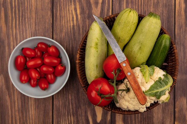 Vue de dessus de légumes frais tels que le concombre courgettes et le chou-fleur sur un seau avec un couteau avec des tomates sur un bol sur un fond en bois