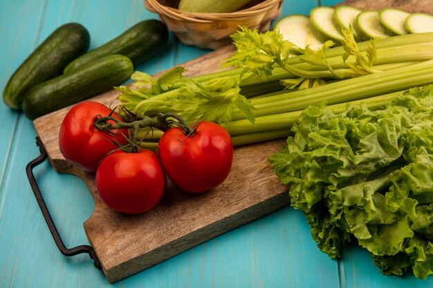 Vue de dessus de légumes frais tels que tomates céleri et courgettes isolés sur une planche de cuisine en bois avec des concombres isolés sur un fond en bois bleu