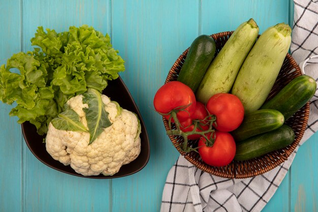 Vue de dessus des légumes frais tels que les tomates, les concombres et les courgettes sur un seau sur un chiffon vérifié avec du chou-fleur et de la laitue sur un bol sur un mur en bois bleu