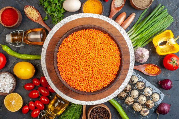 Vue de dessus de lentilles rouges dans un pot marron sur une planche à découper parmi des légumes frais différentes épices oeufs tombés bouteille d'huile sur fond noir