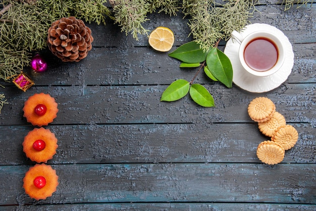 Vue de dessus ligne verticale cupcakes cerises cône sapin feuilles jouets de Noël tranche de citron une tasse de thé et des biscuits sur une table en bois sombre avec espace de copie