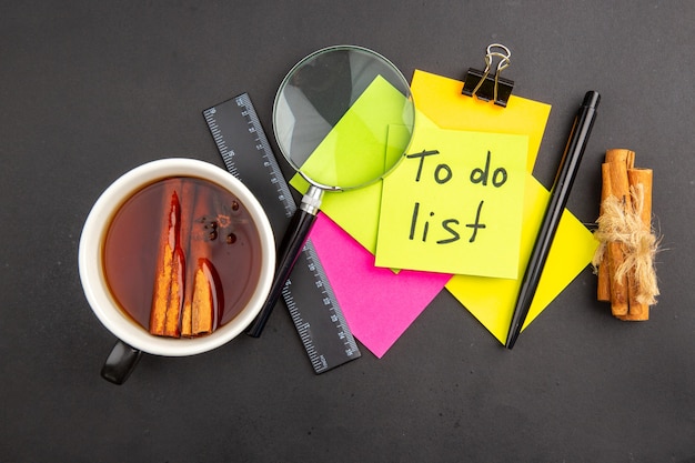 Vue de dessus de la liste de tâches écrite sur une note collante jaune notes collantes colorées stylo lupa bâtons de cannelle règle tasse de thé