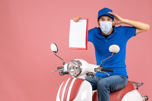 Vue de dessus d'un livreur choqué en masque portant un chapeau assis sur un scooter montrant un document sur une pêche pastel